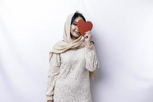 ein glücklich jung asiatisch Muslim Frau tragen ein Hijab fühlt sich romantisch Formen Herz Geste ausdrücken zärtlich Gefühle und halten ein rot herzförmig Papier foto