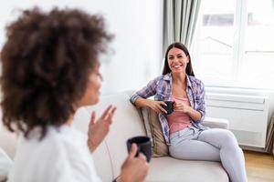 zwei weiblich freunde im Sozial Distanzierung Sitzung auf Sofa im Coronavirus Pandemie Zeit. Beste freunde haben Kaffee zusammen während getrennt durch Sozial Distanzierung auf Sofa beim Zuhause foto