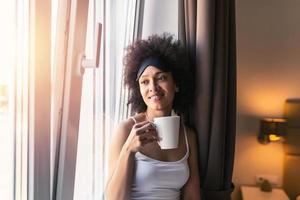 Porträt von lächelnd glücklich heiter schön ziemlich schwarz Frau entspannend Trinken Tasse von heiß Kaffee oder Tee und suchen aus das Fenster. jung Frau mit Schlafen Maske haben Kaffee im das Morgen foto