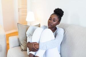 traurig afrikanisch amerikanisch Frau mit Depression Sitzung auf Sofa. einsam betont verärgert jung schwarz Mädchen Denken von psychologisch Problem Denken Bedauern Über Fehler foto