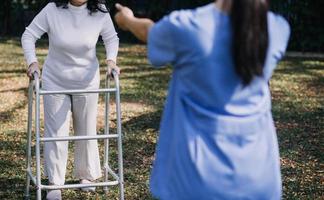 asiatisch jung Pflegekraft Krankenschwester Unterstützung Senior älter männlich Gehen draußen. Spezialist Mädchen Arzt Hilfe und nehmen Pflege von Alten reifen Mann geduldig tun physisch Therapie im Öffentlichkeit Park beim Pflege- heim. foto