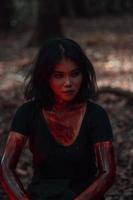 ein asiatisch Frau im schwarz Kleider und bedeckt im Blut war Sitzung im das Mitte von das Wald mit ein unheimlich Gesicht foto