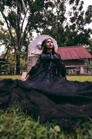 ein jung Hexe im ein lange schwarz Kleid und unheimlich bilden war Sitzung im Vorderseite von das Grabstein foto