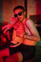 ein Mann mit Sonnenbrille und Nein Kleider posieren mit ein Orange Katze auf ein alt Stuhl foto