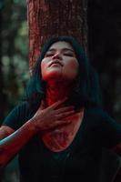 ein asiatisch Frau im schwarz Kleider verschmiert ihr Körper mit rot Blut Vor das Ritual beginnt im Vorderseite von ein groß Baum foto