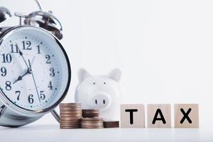 Zeit, Steuern zu zahlen. Sparschweinuhr, gestapelte Münzen und Holzblock auf weißem Hintergrund. Besteuerung und jährliches Steuerkonzept foto