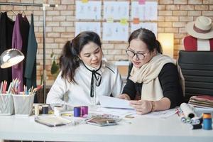 asiatische modedesignerin mittleren alters unterrichtet junge jugendliche schneiderin im studio, zeichnet skizzenstil und nähen von stoffen für kleiderdesign-kollektionsideen, professionelles boutique-kleinunternehmen. foto