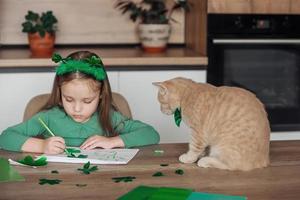 ein wenig Mädchen mit ein Binde auf ihr Kopf zeichnet und schneidet Grün Kleeblätter zum st. Patrick's Tag beim ein Tabelle beim Zuhause im das Küche, Nächster zu ihr ist ihr schön Katze mit ein Grün Bogen Krawatte um seine Hals foto