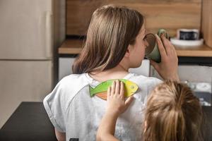 ein fröhliches kleines mädchen klebt ihrer mutter zu hause in der küche einen papierfisch auf den rücken, eine junge frau frühstückt, in der hand hält sie eine tasse tee. lustige familienwitze, aprilscherzfeier foto