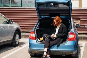 Frau, die am Laptop arbeitet, während sie im Kofferraum des Autos sitzt foto