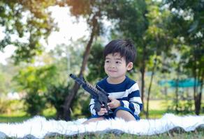 asiatischer Junge, der in einem Park an einem klaren Morgen sitzt und eine Spielzeugpistole hält foto