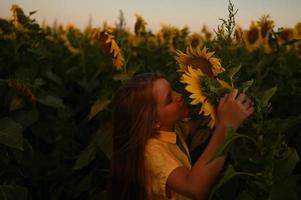 ein glücklich jung Mädchen mit lange Haar im ein Stroh Hut steht im ein groß Feld von Sonnenblumen. Sommer- Tag. ein warm Sonnenuntergang. foto