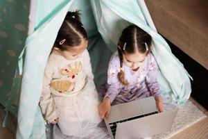 zwei Mädchen Schwestern Aufpassen auf Laptop beim Wigwam Zelt. Technologie und Zuhause Konzept. foto