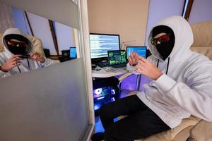 Junge Hacker betrügerisch verwenden Anerkennung Karte zum Zahlung. Internet Diebstahl . Mann tragen ein Sturmhaube und halten ein Anerkennung Karte während saß hinter ein Laptop. foto