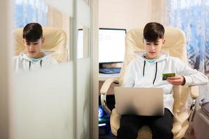 glücklich Teenager Junge mit Laptop und Anerkennung Karte, Sitzung beim Schreibtisch und Bestellung Waren online von heim. foto