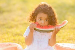 ein wenig dunkel Mädchen mit lockig Haar im ein blass Rosa Kleid isst ein Wassermelone auf das Rasen. Picknick im das Park. glücklich Kindheit. Raum zum Text. hoch Qualität Foto