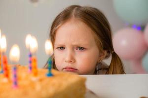 süß wenig Mädchen weht aus Kerzen auf ein Geburtstag Kuchen beim Zuhause gegen ein Hintergrund von Luftballons. Kinder Geburtstag foto