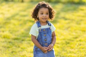 Porträt wenig glücklich spielerisch Kleinkind dunkel Mädchen im ein Denim Sommerkleid Stehen im das Garten auf ein sonnig Tag. Gehen im das frisch Luft. Konzept von ein glücklich Kindheit. Raum zum Text. hoch Qualität Foto