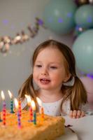 süß wenig Mädchen weht aus Kerzen auf ein Geburtstag Kuchen beim Zuhause gegen ein Hintergrund von Luftballons. Kinder Geburtstag foto