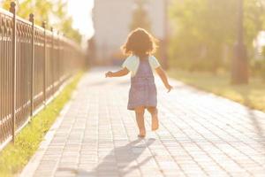 Porträt wenig glücklich spielerisch Kleinkind dunkel Mädchen im ein Denim Sommerkleid Stehen im das Garten auf ein sonnig Tag. Gehen im das frisch Luft. Konzept von ein glücklich Kindheit. Raum zum Text. hoch Qualität Foto