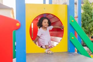 dunkel Baby mit lockig Haar im ein blass Rosa Kleid Theaterstücke auf ein Straße Spielplatz und Fahrten Nieder das hügel. das Konzept von ein gesund Lebensstil. glücklich Kindheit. hoch Qualität Foto