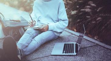 Mann Schreiben Notizbuch mit Laptop im das Park Jahrgang Stil, schlank Hipster Mann im Jeans mit ein Notizbuch, Freiberufler, Kommunikation, Teenager Schüler Junge durchsucht das Internet, Arbeiten draußen das Zuhause foto