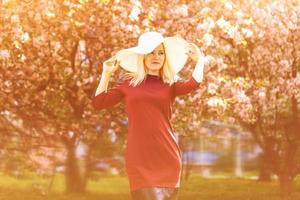 draussen schließen oben Porträt von jung schön Mädchen tragen stilvoll runden Sonnenbrille, modisch violett Hut, Weiß Shirt, posieren im Straße. Rosa Blühen Bäume auf Hintergrund. Frühling Mode Konzept. foto