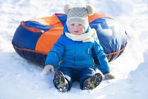 komisch Kind im warm Kleider sitzt im das Schnee mit ein Schläuche. foto