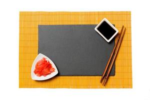 leere rechteckige schwarze Schieferplatte mit Essstäbchen für Sushi, Ingwer und Sojasauce auf gelbem Bambusmattenhintergrund. draufsicht mit kopierraum für ihr design foto