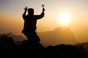 Silhouette von jung Mann beten und halten Christian Kreuz zum Anbetung Gott beim Sonnenuntergang Hintergrund. Christian Religion Konzept Hintergrund. foto