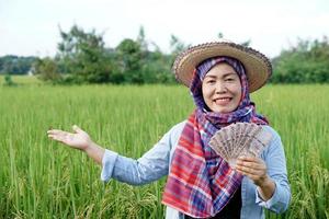 asiatisch Farmer Frau ist beim Paddy Feld, trägt Hut und rot Plaid Shirt, halt thailändisch Banknote Geld. Konzept , Farmer glücklich zu erhalten profitieren, Einkommen, Landwirtschaft unterstützen Geld. foto