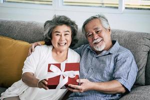 älteres Ehepaar überrascht mit Geschenkbox im Wohnzimmer