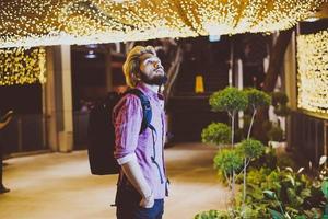 hübscher bärtiger Mann mit Rucksack, der auf der Straße steht, während er nachts reist foto