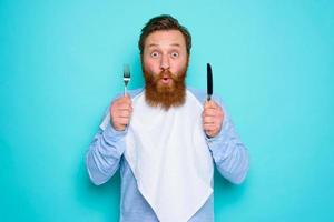 schockiert Mann mit Tätowierungen ist bereit zu Essen etwas mit Besteck im Hand foto