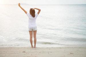 Hinten der jungen Frau stehend strecken ihre Arme in der Luft am Strand mit nackten Füßen foto