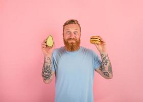 glücklich Mann mit Bart und Tätowierungen ist unentschieden wenn zu Essen ein Avocado oder ein Hamburger foto