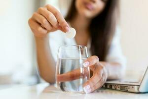 Nahansicht von ein jung Frau fallen lassen ein Brausetabletten Antazida im ein Glas von Wasser. jung Frau kaum stellen ein löslich Pille mit ein Medizin zum Schmerzen oder ein Kater im ein Glas von Wasser foto