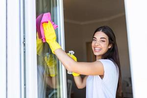 Hausarbeit und Hauswirtschaft Konzept - - glücklich Frau im Handschuhe Reinigung Fenster mit Lappen und Reinigungsmittel sprühen beim Zuhause foto