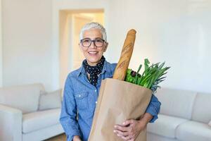 glücklich reifen Frau halten ein Papier Tasche voll von Lebensmittel von das Supermarkt. Alten Frau mit Lebensmittel beim Zuhause foto