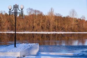 ein Fluss Steg im Winter. Schnee Bank mit Bäume. das Laterne und Seebrücke mit ein Leiter unter das Schnee im das sonnenlicht.foto von hoch Qualität. horizontal foto