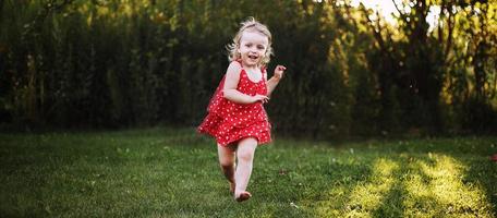 glücklich Baby lächelnd. wenig Mädchen Laufen im das Garten beim Sonnenuntergang draussen barfuß foto