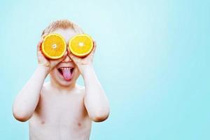 Kind mit Hälften von Orangen auf Augen. glücklich Kind haben Spaß und zeigen Zunge. das Junge ist halten Obst auf ein Licht Blau Hintergrund foto