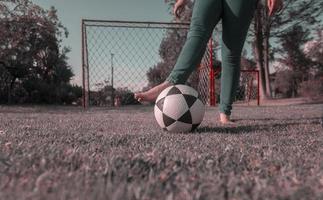 Beine von Frau im Blau Hose, barfuß im Mitte von Park spielen mit ein Fußball Ball mit ein Fußball Tor mit Netze und Bäume im Hintergrund während das Morgen foto