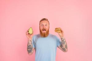 erstaunt Mann mit Bart und Tätowierungen ist unentschieden wenn zu Essen ein Avocado oder ein Hamburger foto