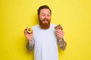 glücklich Mann mit Bart und Tätowierungen hält Avocado und Schokolade foto