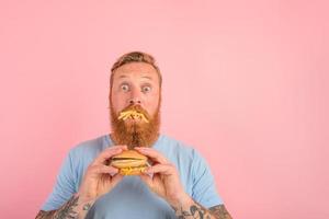 erstaunt Mann mit Bart und Tätowierungen isst ein Sandschalter mit Hamburger und Kartoffeln foto