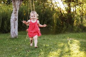 glücklich Baby lächelnd. wenig Mädchen Laufen im das Garten beim Sonnenuntergang draussen barfuß foto