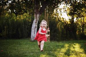 glücklich Baby lächelnd. wenig Mädchen Laufen im das Garten beim Sonnenuntergang draussen barfuß foto