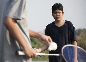 asiatisch Jungs halt Badminton Federball und Schläger, Stehen und spielen neben das Fluss Bank im ihr lokal Fluss während ihr Wochenende Urlaub, Sanft und selektiv Fokus auf Vorderseite Junge im Weiß Shirt. foto