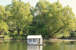 Weißes schwimmendes Entenhaus aus Holz in der Mitte des Teiches im öffentlichen Park. Vogelschutzgebiet im künstlichen See. Frühlingsnest. Sommergrüne Aussicht. Wildlife Naturschutzgebiet im Flusswasser. Unterschlupf für Wasservögel foto